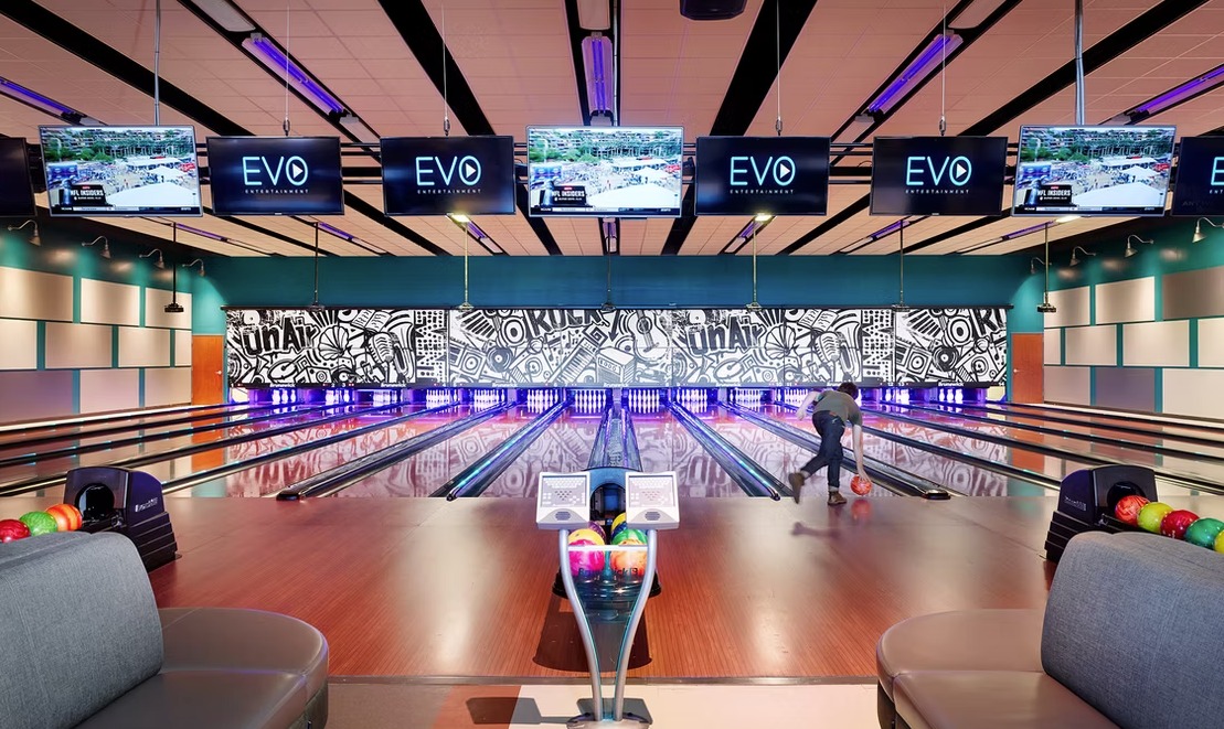 Modern bowling alley featuring multiple lanes with polished wood floors and digital "EVO" displays above each lane. A person is shown mid-motion, rolling a bowling ball.