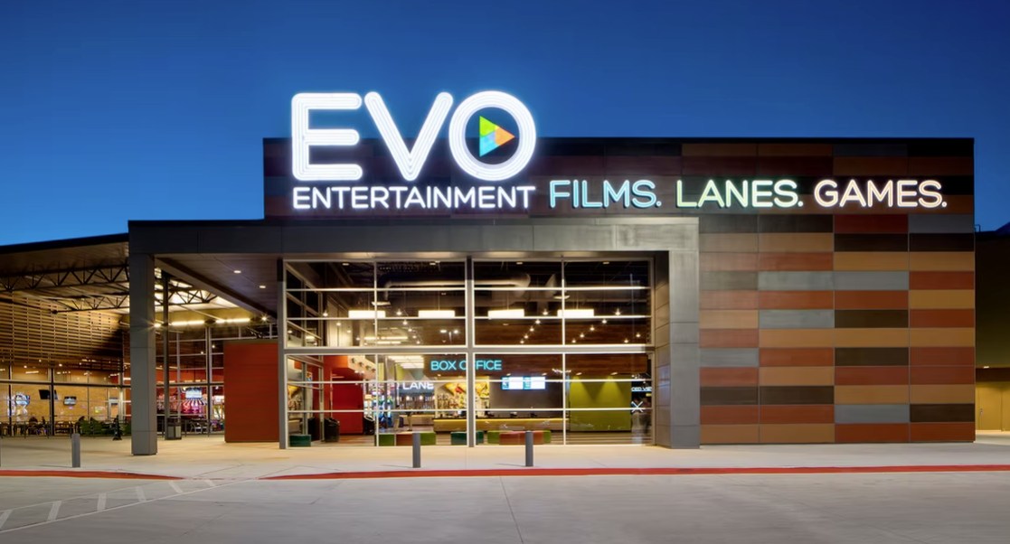 Nighttime view of the EVO Entertainment entrance with a neon sign that reads "FILMS. LANES. GAMES." Behind the large glass windows is a brightly lit lobby.