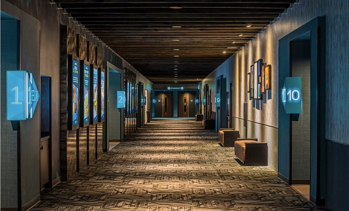 A long, modern-looking hallway for the world's largest movie screen and glowing blue numbered signs marking theater entrances. Digital movie posters are on the walls. .
