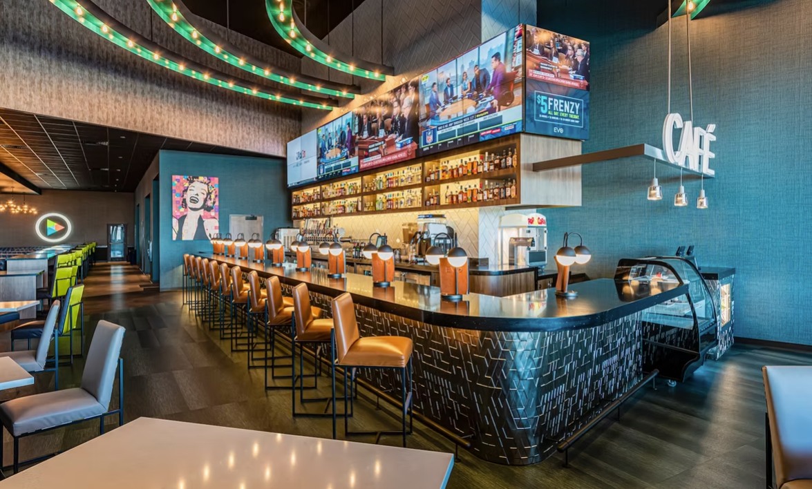 Stylish bar area with a long counter, tan barstools, and large-screen televisions. Curved neon lights decorate the ceiling above. 