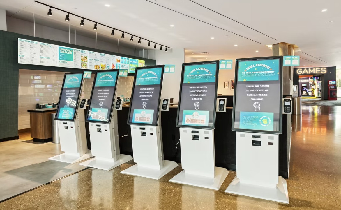 Row of five self-service ticketing kiosks with large touchscreens.. The screens display a welcome message for EVO Entertainment.