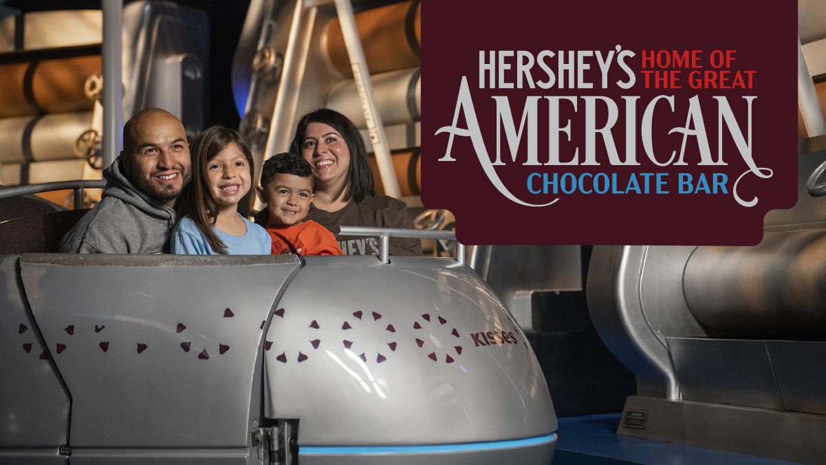 A smiling family of four sitting in a silver ride vehicle decorated with Hershey's Kisses graphics on the Chocolate Tour at Hershey's Chocolate World, with a "Hershey's Home of the Great American Chocolate Bar" logo overlaid in the corner.