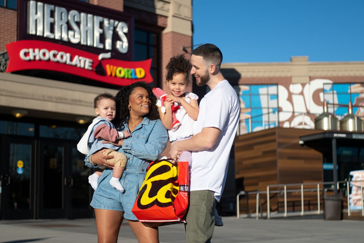 A family of four standing outside the main entrance of Hershey's Chocolate World, with the parents holding a large Reese’s shopping bag and their young children.