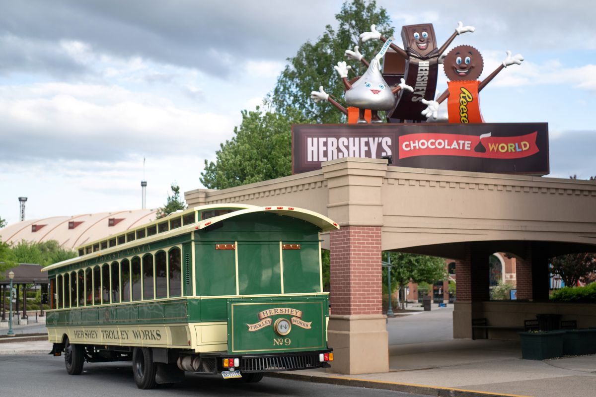 An exterior view of a green and gold Hershey Trolley Works trolley parked in front of the main entrance sign at Hershey's Chocolate World, featuring a giant Hershey’s Kiss and Reese’s characters.