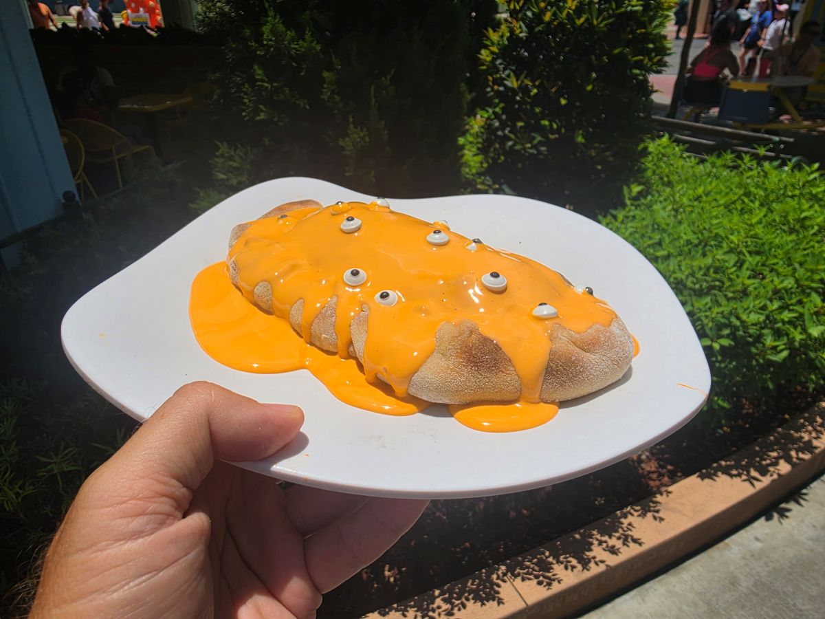 A close-up shot of a person's hand holding a white plate outdoors at Universal Studios Florida, featuring Irene's Creamsicle Cheesecake Calzone from the Minion Cafe. The sweet calzone is decorated with multiple small candy eyeballs scattered across the top.