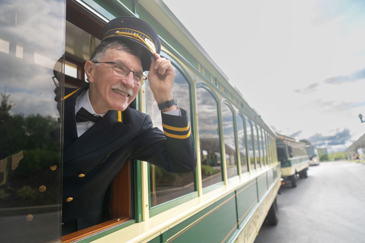 A trolley conductor in a black uniform and cap smiling and waving from the window of a Hershey Trolley Works vehicle.