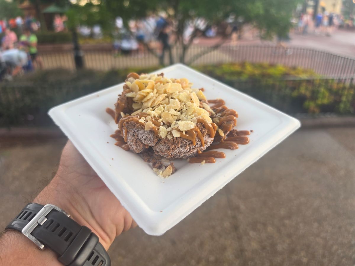 A hand holds a square white plate with a chocolate sweet treat topped with caramel drizzle and almonds set against a blurred park backdrop.