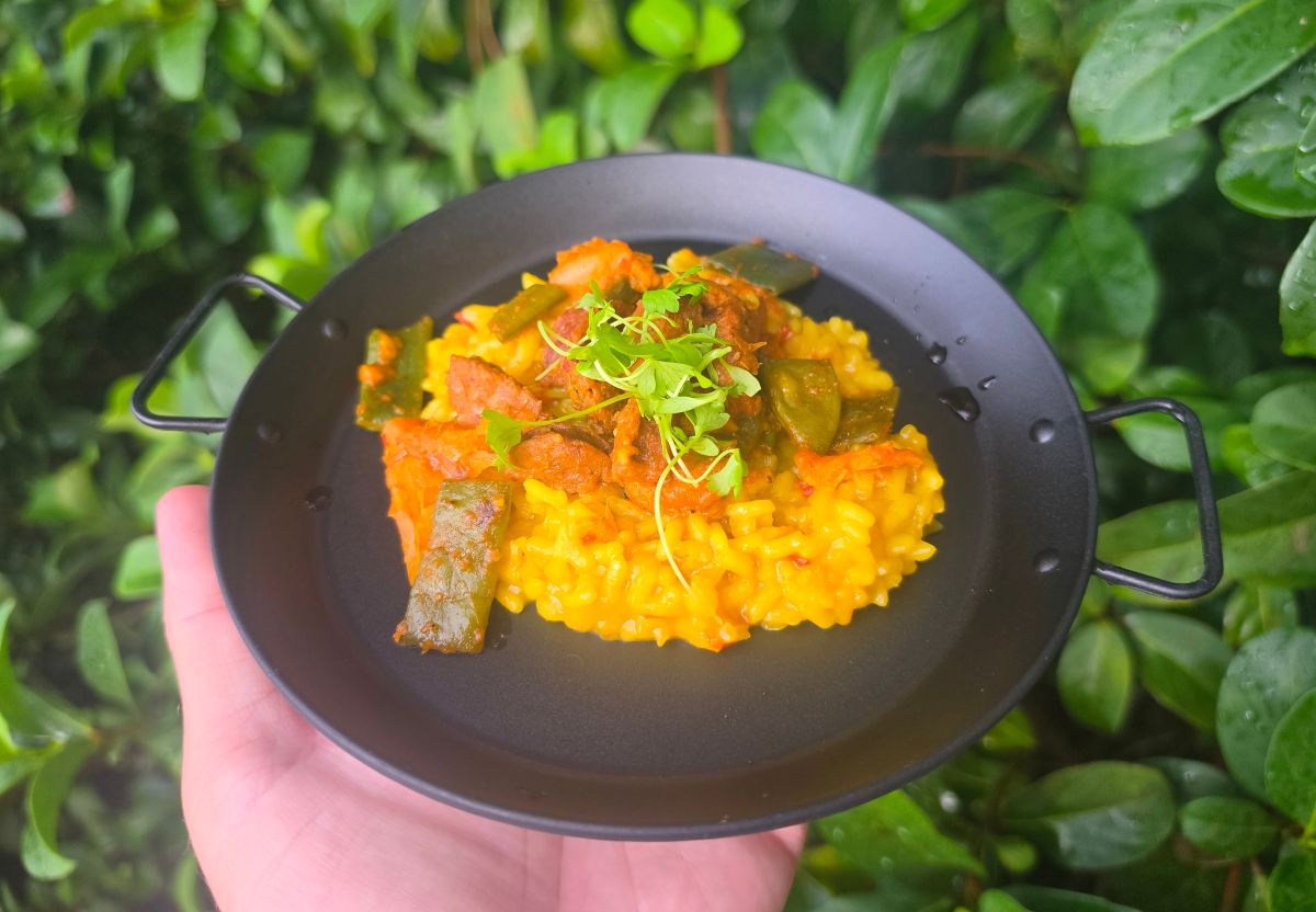 A hand holds a small black pan containing yellow saffron risotto topped with green beans, pieces of meat, and a garnish of fresh green herbs.