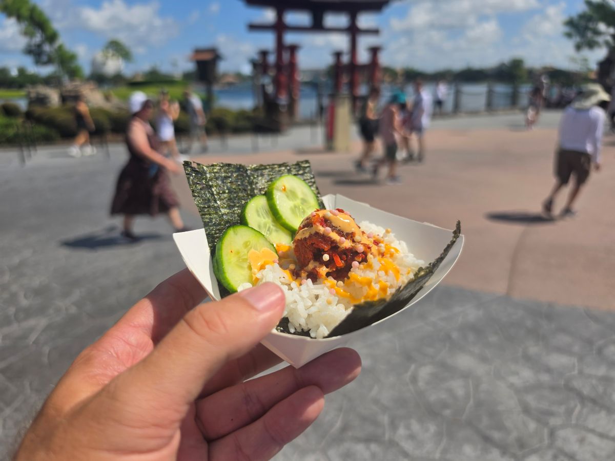 A hand holds a small paper tray with a sushi hand roll filled with rice, cucumber slices, spicy tuna, and sauce, with a Japanese torii gate in the background.