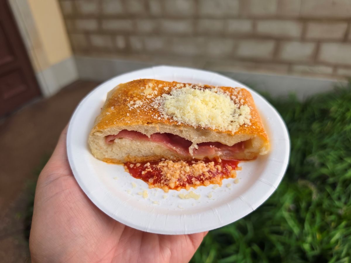 A hand holds a white paper plate with a slice of golden-brown stuffed bread filled with ham, topped with grated cheese and served with red sauce.