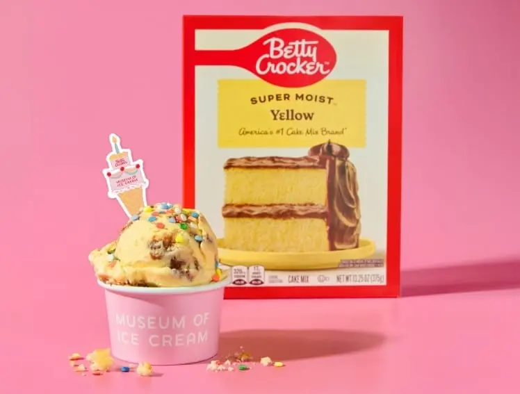 A scoop of yellow cake-flavored Betty Crocker ice cream in a pink Museum of Ice Cream cup, placed in front of a Betty Crocker yellow cake mix box on pink.