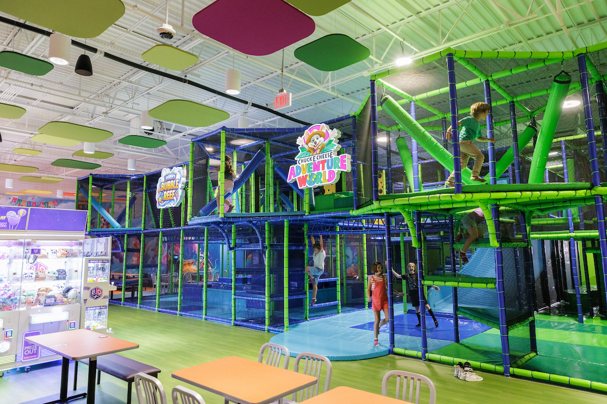 Children play in a large indoor Chuck E. Cheese Adventure World jungle gym with green and blue structures, surrounded by tables and arcade games.