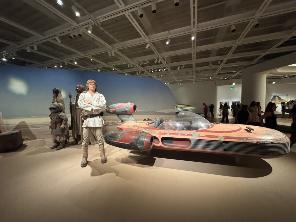 A mannequin of Luke Skywalker stands next to a Landspeeder on display.