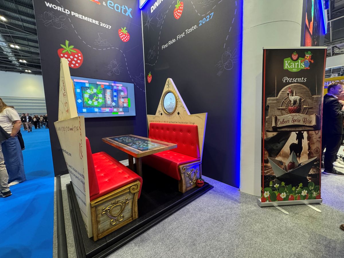 MACK Rides FlowDiner seating vehicle booth display with red cushioned seats and interactive table top on show floor at IAAPA Europe.