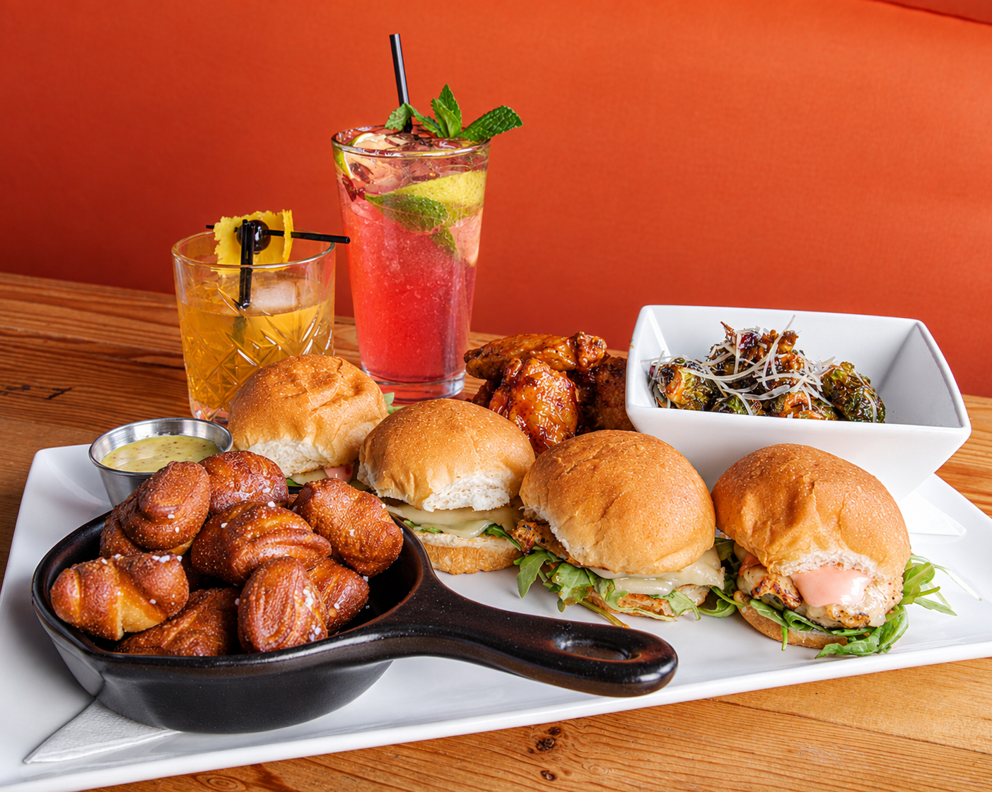 Platter of sliders, pretzel bites in a skillet, chicken wings, roasted Brussels sprouts, and two craft cocktails
