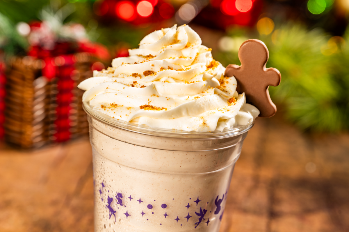 A clear plastic cup filled with a creamy beige beverage, topped with whipped cream, cookie crumbles, and a small gingerbread man-shaped chocolate.