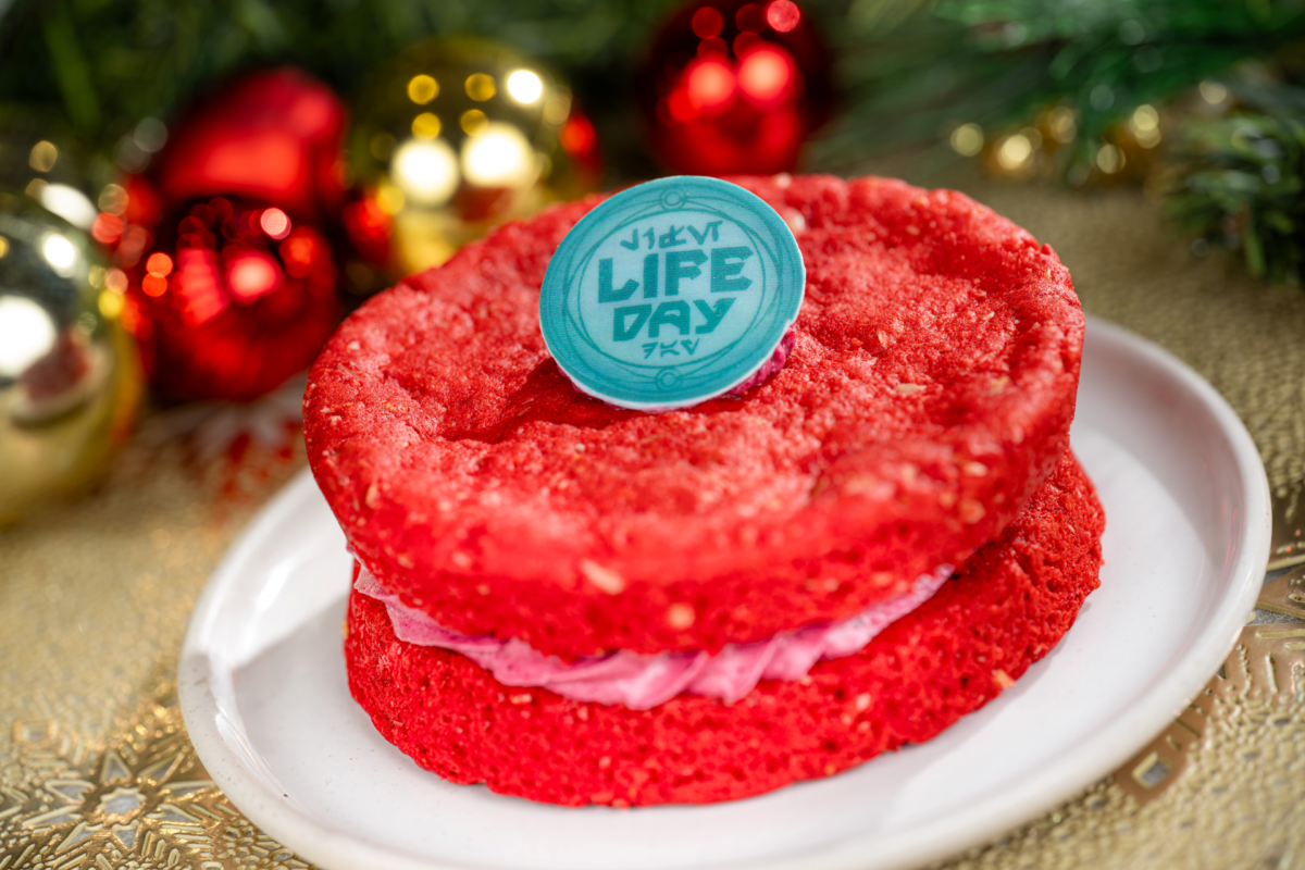 A red sandwich cookie with pink filling on a white plate, topped with a teal Life Day medallion, set against blurred red and gold holiday ornaments.
