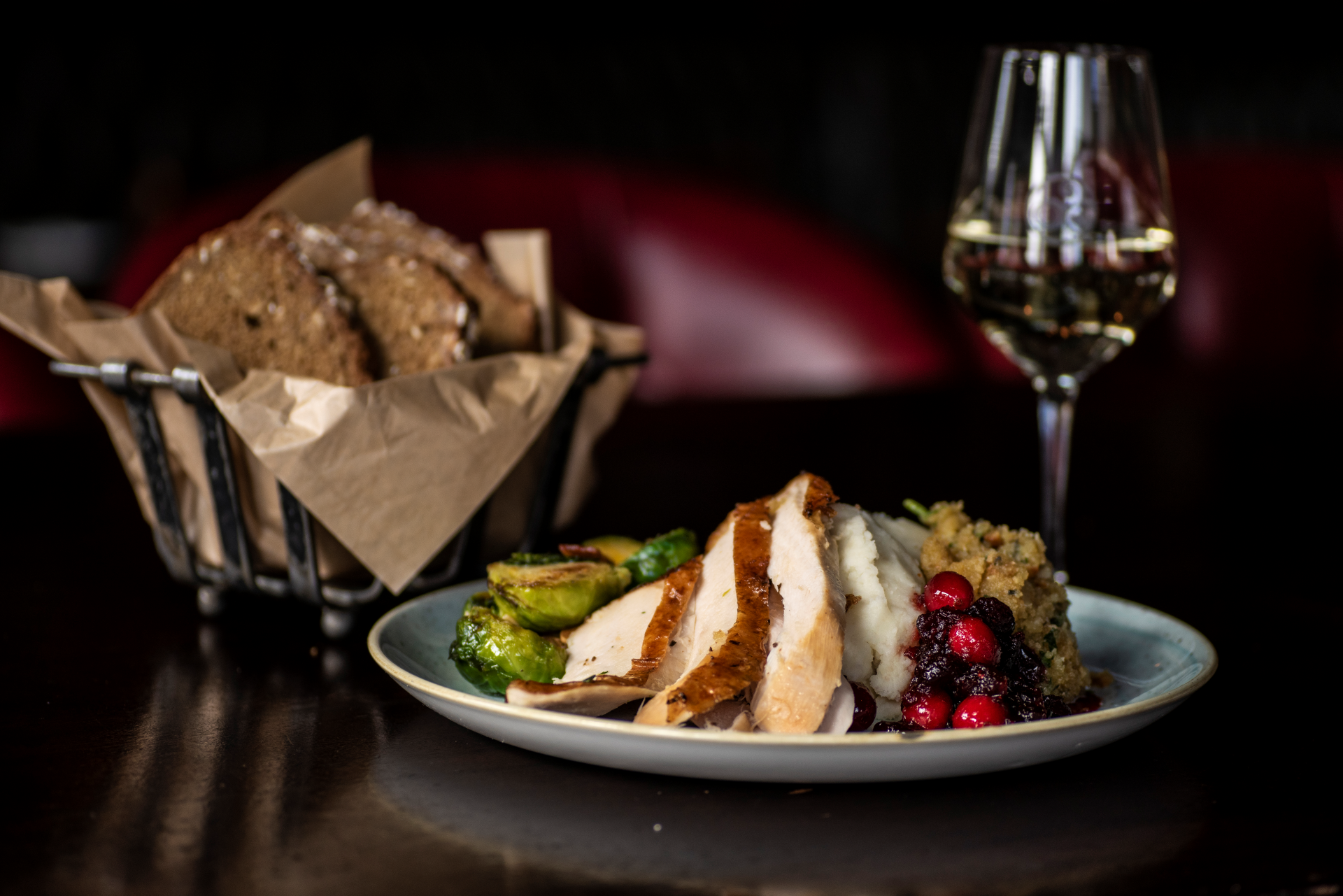 Sliced turkey breast dinner plate with mashed potatoes, stuffing, cranberry sauce, roasted Brussels sprouts, bread basket, and white wine